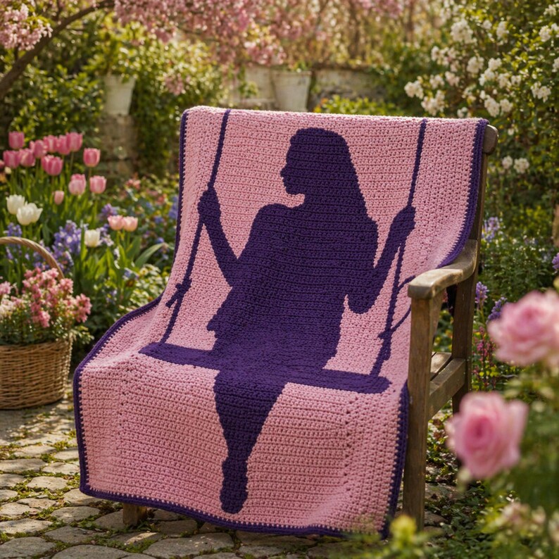 Puede incluir: Una manta de ganchillo rosa y morada sobre un banco de madera. La manta presenta una silueta de una persona en un columpio. El fondo incluye un jard&iacute;n con flores y vegetaci&oacute;n.