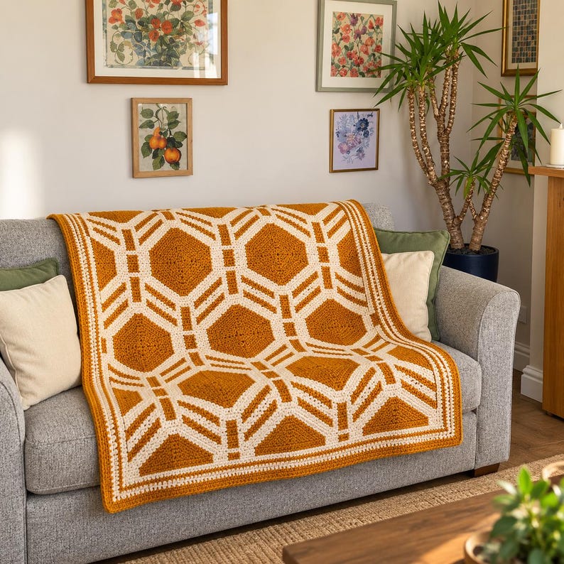 May include: A rectangular crochet blanket featuring a geometric pattern of mustard yellow hexagons outlined in cream white yarn. The blanket has a thick cream white border and is draped over a gray sofa with two solid cream white throw pillows.