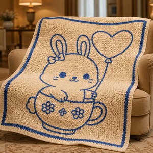 May include: A cream-colored crochet blanket featuring a blue outline of a bunny holding a heart-shaped balloon. The bunny is sitting in a teacup with flower details. The blanket has a blue border and is draped over a beige armchair.