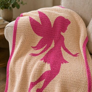 May include: A crocheted blanket in beige with a large pink fairy silhouette. The blanket has a pink border and is draped over a sofa. The fairy design features outstretched arms and a flowing skirt.