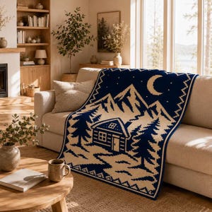 May include: A rectangular knit throw blanket featuring a dark blue and cream color scheme. The design depicts a cabin nestled among pine trees and mountains under a crescent moon and stars, finished with a geometric border pattern.
