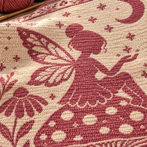 May include: A cream-colored crochet blanket featuring a dark pink tapestry design of a fairy sitting on a large mushroom. The pattern includes a crescent moon, stars, and floral elements. The texture shows individual crochet stitches throughout.
