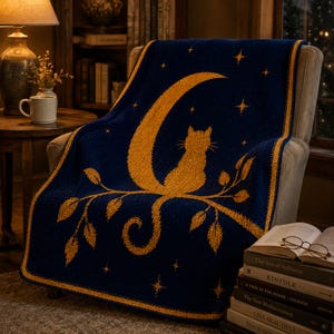 May include: A navy blue knitted throw blanket featuring a gold crescent moon, a cat silhouette, and stars. The blanket is draped over a chair. The blanket's border is gold. The image also shows a stack of books and a lamp.