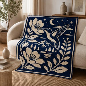 May include: A rectangular woven throw blanket featuring a dark navy blue background with a cream-colored graphic design. The pattern depicts a hummingbird hovering near a large flower, with a crescent moon, stars, mountains, and pine trees.