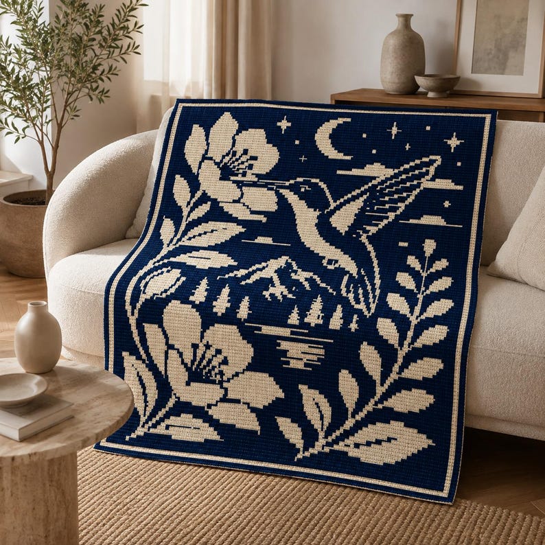 May include: A rectangular woven throw blanket featuring a dark navy blue background with a cream-colored graphic design. The pattern depicts a hummingbird hovering near a large flower, with a crescent moon, stars, mountains, and pine trees.