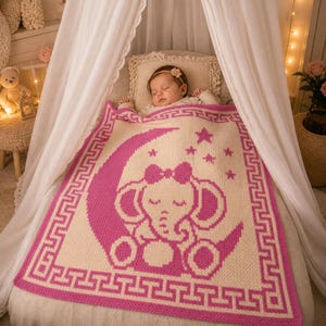 May include: A pink and cream-colored baby blanket with an elephant design. The blanket features a sleeping elephant on a crescent moon, surrounded by stars. The border has a Greek key pattern. The blanket is approximately 40 inches by 40 inches.