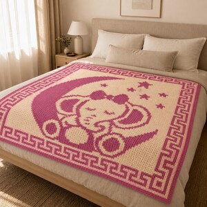 May include: A handmade, square baby blanket featuring a sleeping elephant design. The blanket is primarily cream-colored with a pink elephant, moon, stars, and border. The elephant wears a bow and is surrounded by stars.