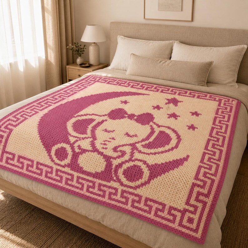 May include: A handmade, square baby blanket featuring a sleeping elephant design. The blanket is primarily cream-colored with a pink elephant, moon, stars, and border. The elephant wears a bow and is surrounded by stars.