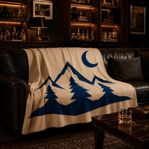 May include: A beige throw blanket featuring a bold, dark blue graphic print of a mountain range, three evergreen trees in the foreground, and a crescent moon in the upper right corner. The blanket is draped over a dark leather sofa.