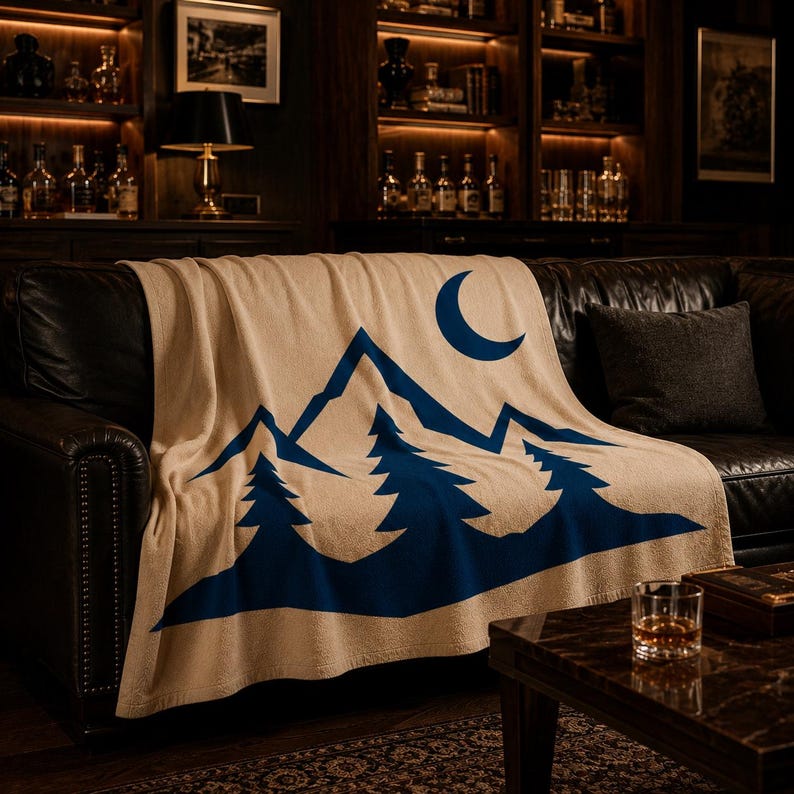 May include: A beige throw blanket featuring a bold, dark blue graphic print of a mountain range, three evergreen trees in the foreground, and a crescent moon in the upper right corner. The blanket is draped over a dark leather sofa.