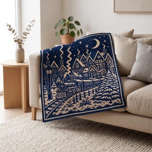 May include: A rectangular knit throw blanket featuring a dark blue and cream color scheme. The design depicts a winter village scene with a steam train on tracks, houses with smoking chimneys, a tall street lamp, a pine tree, and a crescent moon.