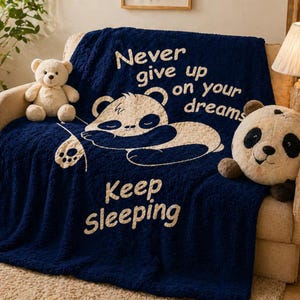 May include: A navy blue fleece blanket with the text "Never give up on your dreams" and "Keep Sleeping" in white. A panda illustration is featured on the blanket. A teddy bear and a panda cushion are on the sofa.