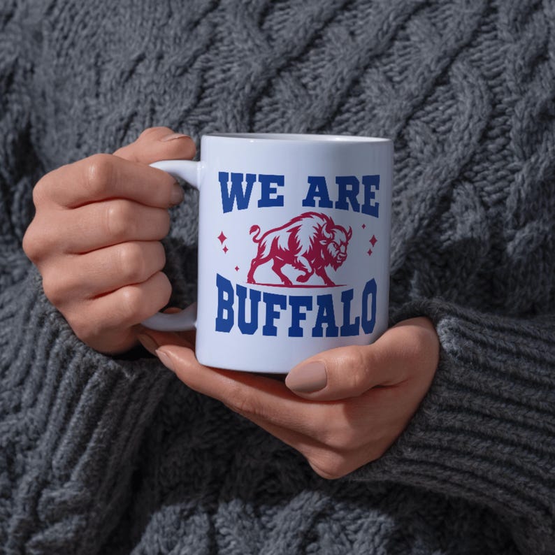 May include: White ceramic mug with the text "WE ARE BUFFALO" in blue, with a red and white bison graphic. The mug has a silver rim and a white handle. The mug is held in front of a gray sweater.