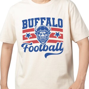 May include: Cream-colored t-shirt featuring a blue and red graphic. The design includes the word "BUFFALO" above "Football", with a football helmet and stars. The shirt is short-sleeved.