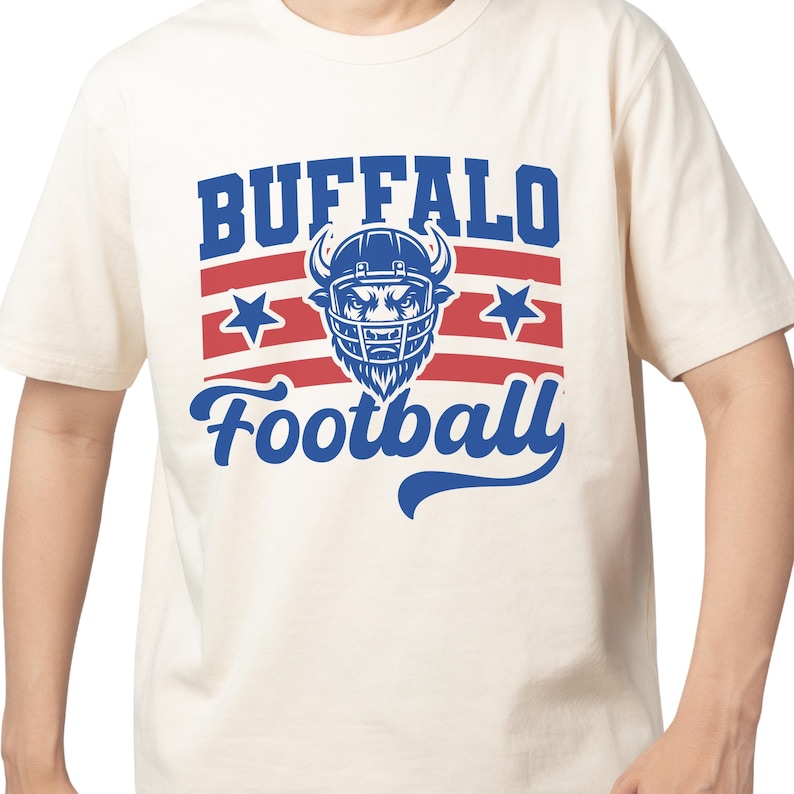 May include: Cream-colored t-shirt featuring a blue and red graphic. The design includes the word "BUFFALO" above "Football", with a football helmet and stars. The shirt is short-sleeved.