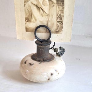 May include: A vintage-style photo holder featuring a round, speckled off-white stone base with a dark metal neck and a circular wire clip. A black and white photograph of a person wearing an aviator cap and goggles is held in the wire clip.