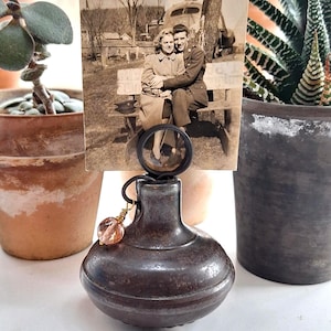 May include: A dark metallic, rounded base photo holder featuring a circular wire clip at the top. A vintage black and white photograph of a couple sitting on a bench is held in the clip. A small, faceted, light pink glass bead hangs from a wire loop.
