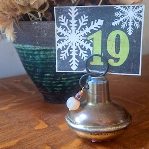 May include: A brass door knob repurposed as a card holder with a dark metal wire loop at the top. A rectangular card with a black background, a white snowflake graphic, and the large green number 19 is inserted. A small bead charm hangs from the wire.