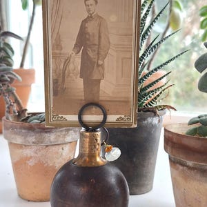 May include: A vintage photo holder featuring a sepia-toned portrait of a man. The holder has a dark brown, spherical base with a brass-colored top and a black ring to hold the photo. A small charm hangs from the ring.