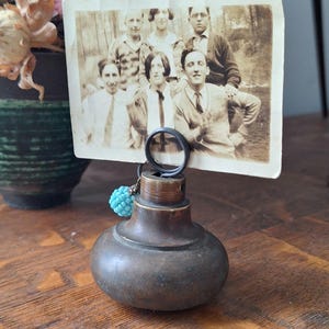 May include: A vintage photo holder with a round, bronze-coloured base and a black metal ring to hold a photograph. A small, turquoise beaded charm hangs from the ring. The photo shows a group of people in sepia tones.