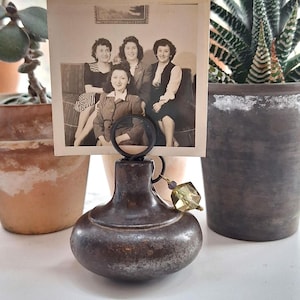 May include: A vintage photo holder with a dark, aged metal finish. The holder has a round base and a loop to hold a photograph. A black and white photograph of four women is displayed. A decorative green crystal hangs from the holder.
