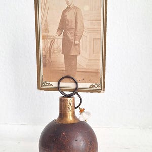 Peut inclure: Un porte-photo vintage avec un portrait sépia d'un homme en manteau sombre. Le support a une base sphérique marron foncé avec un col couleur laiton et un anneau décoratif pour tenir la photo.