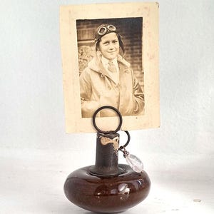 May include: A brown ceramic picture holder with a round base and a narrow neck. A vintage photo of a young boy wearing goggles and a leather jacket is held in place by a metal ring. A clear crystal charm hangs from the ring.