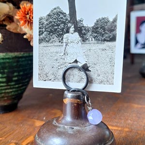May include: A vintage photo holder featuring a black and white photograph of a person sitting under a tree. The holder has a dark brown, bulbous base with a black ring and a light purple bead charm.
