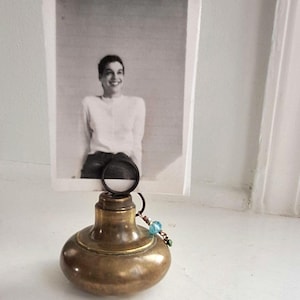 May include: A vintage brass photo holder with a round base and a circular clip for a photograph. The black and white photo shows a smiling person. A small beaded charm hangs from the holder. The holder is likely made in the UK.