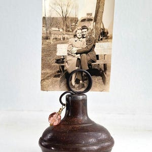 May include: A vintage photo holder featuring a sepia-toned photograph of a couple. The holder has a dark brown, bulbous base with a circular frame to hold the photo. A pink, faceted bead hangs from the frame.