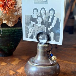 May include: A vintage bronze photo holder with a black ring to hold a black and white photograph. The holder has a decorative base and a small charm with blue and gold beads.