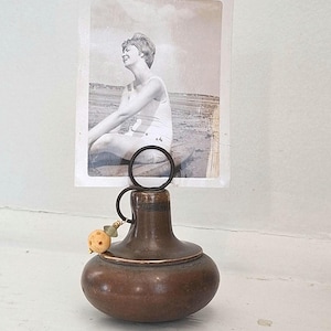 May include: A vintage-style photo holder with a bronze-colored base and a circular wire top. The holder displays a black and white photograph of a person in a swimsuit. A small, decorative bead hangs from the wire.