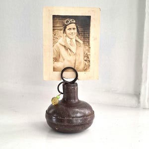 May include: A dark brown, rounded metal base with a circular wire holder at the top, displaying a vintage black and white photograph of a person wearing aviator goggles and a jacket. A small, translucent yellow and green bead charm hangs from the ring.