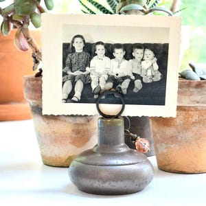 May include: A dark metallic, rounded photo holder base with a circular wire clip at the top, featuring a small dangling charm with a faceted pink bead. A vintage black and white photograph of five children sitting on a sofa is held in the clip.