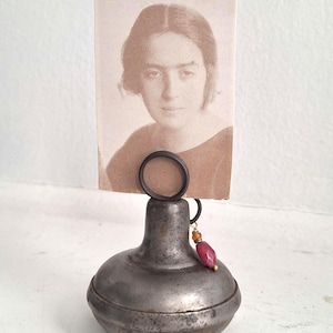 May include: A vintage metal photo holder with a sepia-toned portrait. The holder has a round base and a ring to hold the photo. A small, red gemstone hangs from the ring. The photo shows a woman with short hair.