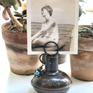 May include: A dark brown, rounded metal object serving as a photo holder, featuring a black wire loop at the top that secures a vintage black and white photograph of a person sitting on a beach. A small blue glass bead hangs from the wire loop.