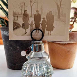 May include: A clear glass photo holder with a silver metal top and a decorative, rounded base. The holder displays a vintage black and white photograph of a family. A small charm with a pink bead and red beads hangs from the top.