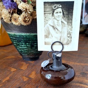 May include: A brown ceramic picture holder with a metal ring and a small, clear stone charm. The holder is holding a black and white photo of a young person wearing a leather jacket and aviator goggles.