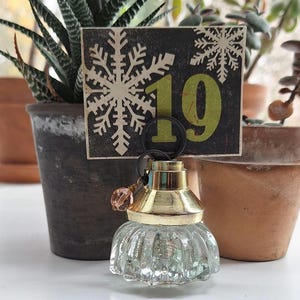 May include: A clear glass bottle stopper with a gold-colored top and a decorative pink bead. A small black tag with the number "19" in green and snowflake designs is behind the bottle stopper.