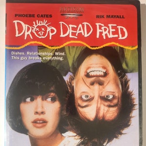 May include: DVD cover for the movie "Drop Dead Fred" starring Phoebe Cates and Rik Mayall. The cover features a red background with the title in yellow and white text. The image shows Cates and Mayall in a close-up shot, with Mayall upside down. The tagline reads "Dishes. Relationships. Wind. This guy breaks everything."