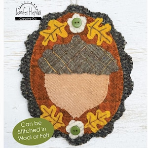 May include: A wool fall acorn ornament featuring a central tan acorn shape with a dark grey textured cap. The design is framed by a rust-coloured border with yellow oak leaves and two white flowers with green buttons. Text reads Wool Fall Acorn Ornament.