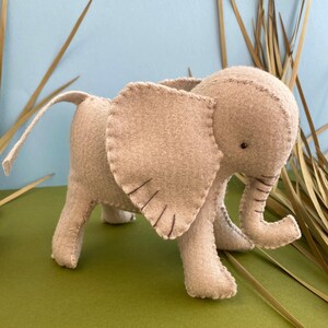 May include: A beige felt elephant toy with stitched details, including ears, trunk, and eyes. The toy is standing on a green surface.