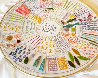 May include: A circular embroidery hoop with a white fabric background. The fabric is divided into 30 sections, each containing a different embroidery stitch and its name. The stitches are embroidered in various colors, including pink, yellow, green, blue, and purple. The text "30 Day Sampler Volume 1" is written in the center of the hoop.