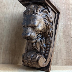 Op de afbeelding: Een gebeeldhouwde houten leeuwenkop console in bruine tinten. Het gezicht van de leeuw is gedetailleerd met een manen, neus en poot. De console heeft een rechthoekige bovenkant en een driehoekige basis.