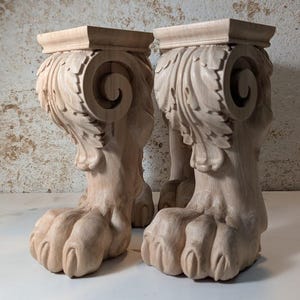 Puede incluir: Dos patas de muebles de madera con intrincados tallados de hojas y volutas. Las patas están acabadas en un color de madera natural y tienen un diseño de pata de garra.