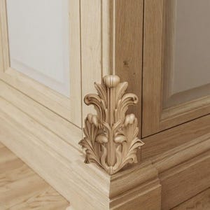 May include: A light-colored wooden cabinet corner featuring a vertical carved acanthus leaf ornament. The wood grain is visible throughout the structure, which includes a base molding and a flat panel door section with clean, straight lines.