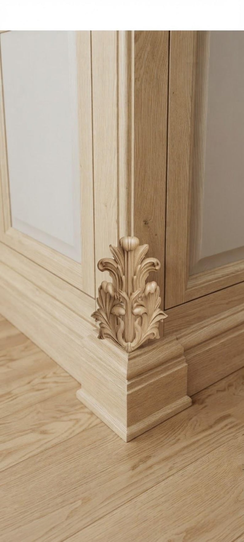 May include: A light-colored wooden cabinet corner featuring a vertical carved acanthus leaf ornament. The wood grain is visible throughout the structure, which includes a base molding and a flat panel door section with clean, straight lines.