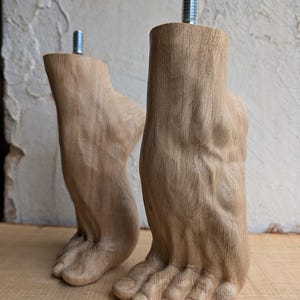 Op de afbeelding: Twee lichtbruine houten meubelpoten uitgesneden in de vorm van menselijke voeten. Elke poot heeft een zichtbare houtnerfstructuur en een metalen draadeind aan de bovenkant voor bevestiging. De voeten staan rechtop op een vlakke ondergrond.