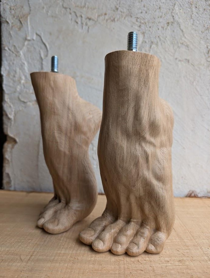Op de afbeelding: Twee lichtbruine houten meubelpoten uitgesneden in de vorm van menselijke voeten. Elke poot heeft een zichtbare houtnerfstructuur en een metalen draadeind aan de bovenkant voor bevestiging. De voeten staan rechtop op een vlakke ondergrond.