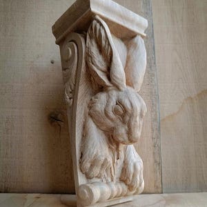 May include: A wooden corbel with a carved rabbit design. The corbel is rectangular, with a decorative top and bottom. The rabbit is carved in detail. The wood is a natural, light brown colour, and the corbel is suitable for interior decor.